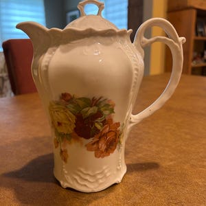 May include: White porcelain teapot with a lid and handle. The teapot is decorated with a floral design featuring yellow and orange roses. The teapot has a curved spout and a decorative handle.