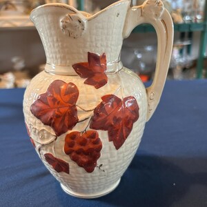 May include: Cream-colored ceramic pitcher with a textured basketweave pattern and a decorative handle. The pitcher is adorned with raised, reddish-brown grape leaves and clusters, suggesting a vineyard theme. A classic piece for serving beverages.