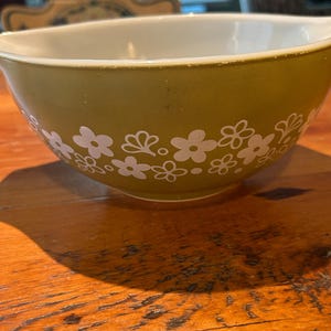May include: A round mixing bowl with a smooth olive green exterior and a white interior. The bowl features a repeating white floral pattern around the center, consisting of various stylized flower shapes and small dots on the green surface.