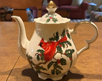 May include: White ceramic teapot with a gold handle and accents. The teapot features a red cardinal bird design with green holly leaves and red berries. The lid is topped with a gold finial. A festive holiday themed teapot.
