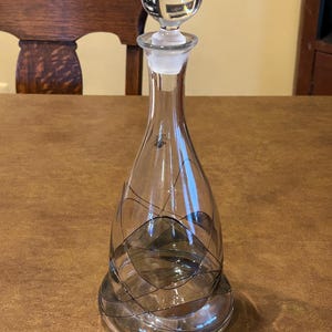 May include: A clear glass decanter with a spherical stopper. The decanter has a teardrop shape and is decorated with a black and gray abstract design. The stopper is clear and spherical. The decanter is likely used for serving wine or other beverages.