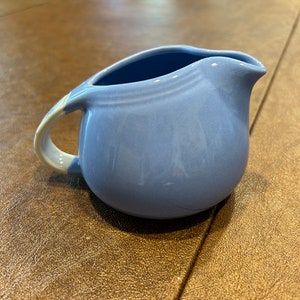 May include: A small, light blue ceramic creamer with a white handle. The creamer has a rounded shape and a slightly flared lip.
