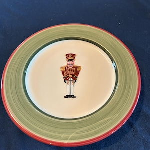 May include: A round decorative plate with a green and red rim. The center of the plate features a detailed illustration of a toy soldier in a red and gold uniform, a black hat, and white pants. The plate is on a dark blue surface.