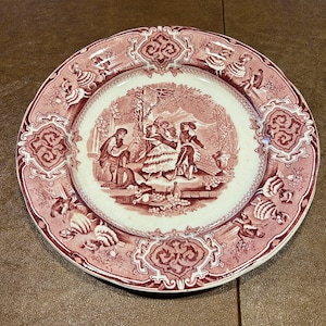 May include: A vintage pink and white ceramic plate with a floral border and a central scene depicting a group of people in a garden setting.