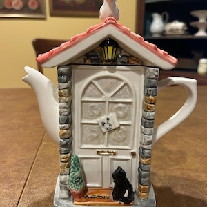 May include: A whimsical ceramic teapot shaped like a cottage. The teapot features a white door with a "Back 4 Tea" sign, a black cat, and a welcome mat. A bird sits atop the red-tiled roof. The teapot has a white spout and handle.