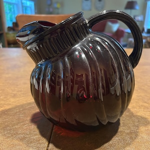 May include: A dark red glass pitcher with a ribbed, pumpkin-shaped body and a curved handle. The pitcher is sitting on a brown surface. The pitcher is likely used for serving beverages.