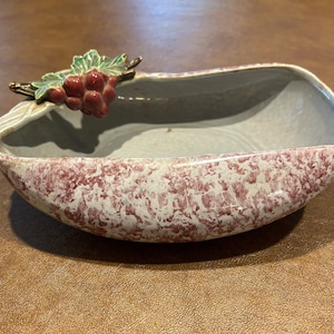 May include: A white ceramic planter with a red and white speckled glaze. The planter is shaped like a grape leaf and has a grape cluster attached to the side.