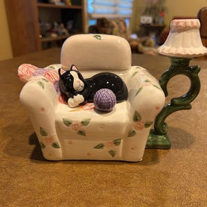 May include: A ceramic figurine of a cream-colored armchair with a floral pattern, featuring a sleeping black and white cat, a purple ball of yarn, and an attached green lamp with a pink-trimmed shade extending from the right side.