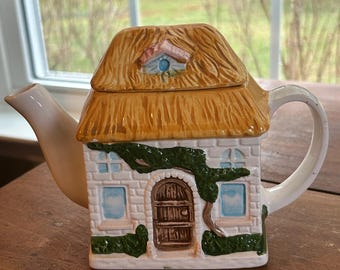 May include: A ceramic cottage-shaped teapot. The roof is straw-coloured, and the walls are white brick with a brown door and green vines. The teapot has a white spout and handle, and a small window on the roof.