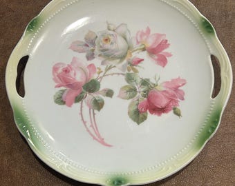 Könnte beinhalten: Ein rundes Serviertablett aus weißem Porzellan mit zwei integrierten Seitengriffen. Die Mitte zeigt eine florale Illustration aus rosa und weißen Rosen mit grünen Blättern. Der gewellte Rand ist mit einer blassgelben Umrandung akzentuiert.