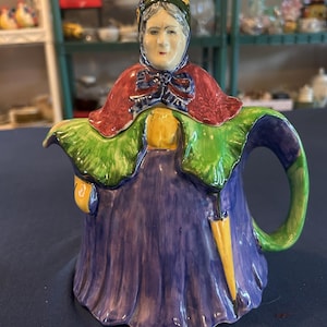 May include: A ceramic teapot in the shape of an elderly woman. The teapot features a purple dress, a red shawl, and a green and blue bonnet. The handle is green, and a yellow umbrella is visible. The teapot is painted with bright colors.