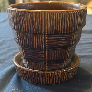 May include: A dark brown ceramic plant pot with a matching saucer. The exterior features a textured, basket-weave pattern with alternating vertical and horizontal grooved lines, creating a grid-like appearance around the entire vessel.
