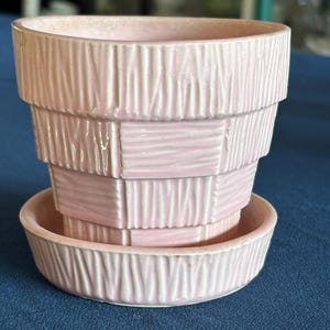 May include: A light pink ceramic planter with a tiered, stepped silhouette and a matching circular saucer. The exterior surface features a textured pattern of vertical and horizontal lines, creating a woven or basket-like appearance in a glossy finish.