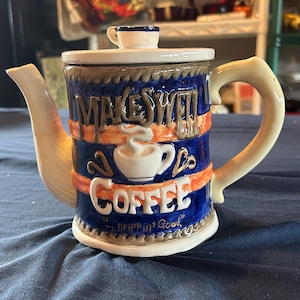 May include: A ceramic teapot with a blue body, cream-colored handle and spout, and a lid featuring a miniature cup handle. The front displays the embossed text "MAKES WELL" and "COFFEE" with a cup graphic and the phrase "Drippin' Good".