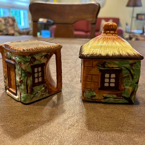 Puede incluir: Un juego de azucarera y jarra de cerámica. La jarra tiene un asa y la azucarera tiene tapa. Ambos tienen forma de casitas con tejados de paja, paredes de ladrillo y pequeñas ventanas. En las paredes hay enredaderas y flores verdes.