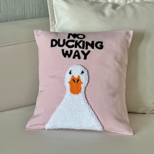 Op de afbeelding: Een roze sierkussen met de tekst "NO DUCKING WAY" in zwart boven een wit ganzenontwerp met een oranje snavel. Het kussen ligt op een witte bank met een wit kussen erachter.