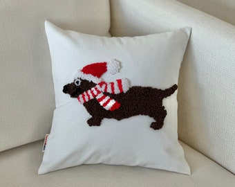 Può includere: Cuscino bianco con un bassotto marrone che indossa una sciarpa a righe rosso e bianco e un cappello di Babbo Natale rosso.
