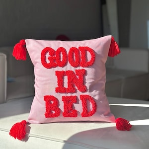 Peut inclure: Un coussin rose avec les mots "GOOD IN BED" brodés en rouge, orné de pompons rouges à chaque coin. Le coussin est posé sur un canapé blanc, créant une ambiance chaleureuse et accueillante.