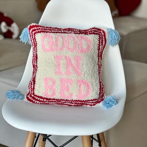 Peut inclure: Un oreiller blanc avec une bordure rouge et blanche et des glands bleus. L'oreiller est fait d'un matériau moelleux et porte les mots "Good in Bed" écrits en lettres roses.