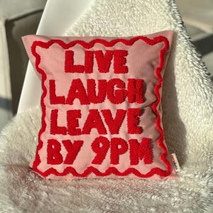 Può includere: Cuscino rosa con un messaggio ricamato in rosso che recita "Live Laugh Leave By 9PM".