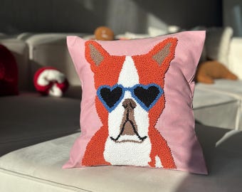 May include: A pink square decorative pillow featuring a dog design. The dog has orange fur, white markings, and wears heart-shaped blue sunglasses. The pillow is placed on a white sofa.
