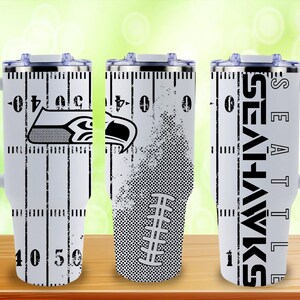 Puede incluir: Vaso t&eacute;rmico blanco con un dise&ntilde;o de campo de f&uacute;tbol, el logotipo y el nombre del equipo de los Seattle Seahawks. El vaso tiene un asa y una tapa. El dise&ntilde;o incluye l&iacute;neas de yardas y un gr&aacute;fico de f&uacute;tbol americano.