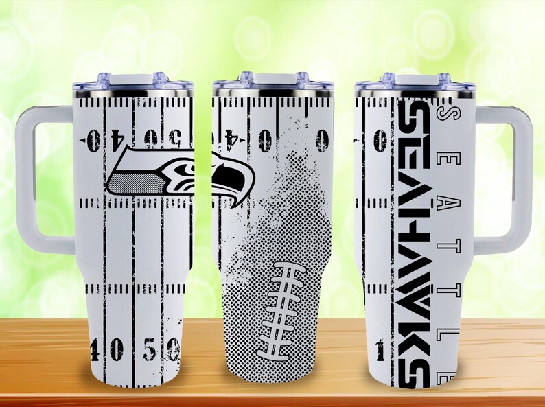 Puede incluir: Vaso t&eacute;rmico blanco con un dise&ntilde;o de campo de f&uacute;tbol, el logotipo y el nombre del equipo de los Seattle Seahawks. El vaso tiene un asa y una tapa. El dise&ntilde;o incluye l&iacute;neas de yardas y un gr&aacute;fico de f&uacute;tbol americano.