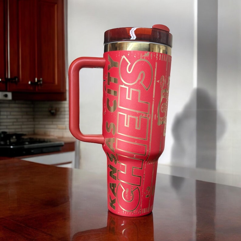 K&ouml;nnte beinhalten: Ein roter Reisebecher mit Henkel, mit dem goldenen Schriftzug "KANSAS CITY CHIEFS". Der Becher hat einen schwarzen und goldenen Deckel und ist so konzipiert, dass er Getr&auml;nke hei&szlig; oder kalt h&auml;lt. Der Becher steht auf einer Holzoberfl&auml;che.