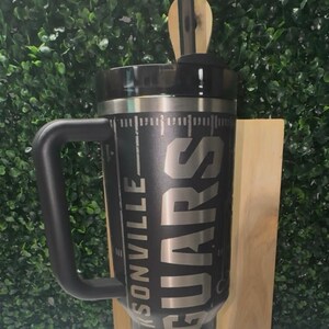 Puede incluir: Vaso t&eacute;rmico negro con asa, con el texto "JACKSONVILLE JAGUARS" en plata. El vaso tiene una tapa negra y una pajita. El dise&ntilde;o incluye un gr&aacute;fico similar a un campo de f&uacute;tbol. Un regalo perfecto para cualquier aficionado al f&uacute;tbol.