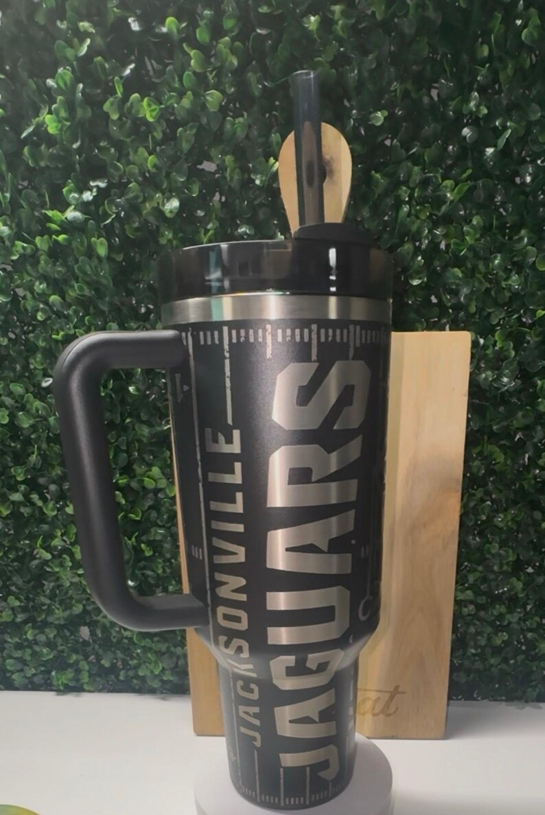 Puede incluir: Vaso t&eacute;rmico negro con asa, con el texto "JACKSONVILLE JAGUARS" en plata. El vaso tiene una tapa negra y una pajita. El dise&ntilde;o incluye un gr&aacute;fico similar a un campo de f&uacute;tbol. Un regalo perfecto para cualquier aficionado al f&uacute;tbol.