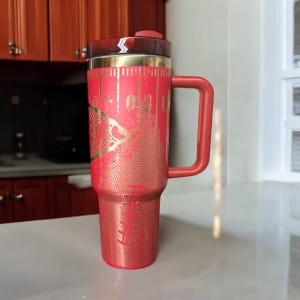K&ouml;nnte beinhalten: Ein roter isolierter Reisebecher mit Henkel und Deckel. Der Becher hat ein goldfarbenes Design und Text. Der Griff und der Deckel sind rot. Der Becher ist so konzipiert, dass er Getr&auml;nke hei&szlig; oder kalt h&auml;lt.