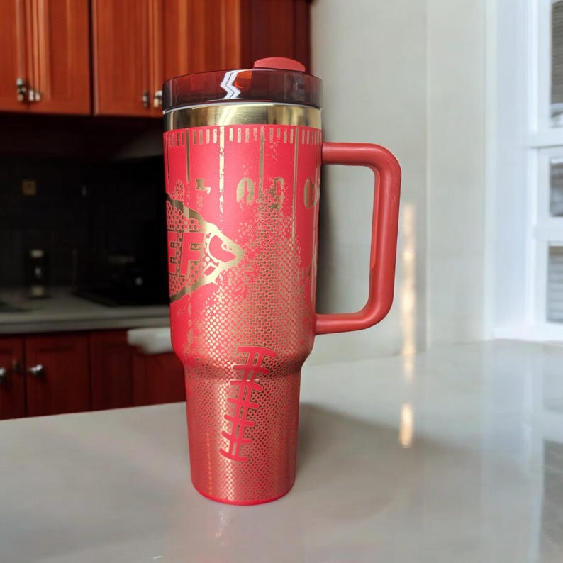 K&ouml;nnte beinhalten: Ein roter isolierter Reisebecher mit Henkel und Deckel. Der Becher hat ein goldfarbenes Design und Text. Der Griff und der Deckel sind rot. Der Becher ist so konzipiert, dass er Getr&auml;nke hei&szlig; oder kalt h&auml;lt.