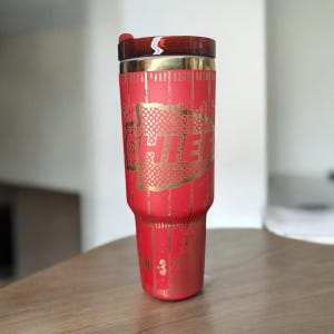 K&ouml;nnte beinhalten: Ein roter Isolierbecher mit goldenen Akzenten und dem Wort "CHIEFS" in einem goldenen, gepunkteten Design. Der Deckel ist braun mit einem wei&szlig;en und roten Streifen. Der Becher hat ein Fassungsverm&ouml;gen von ca. 887 ml.