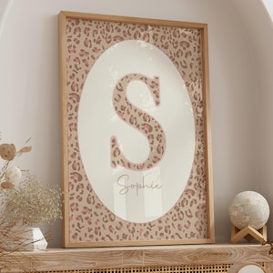 Könnte beinhalten: Ein gerahmter Druck mit einem großen Buchstaben "S" im Leopardenmuster, platziert in einem ovalen Feld auf hellem Hintergrund. Der Name "Sophie" steht darunter in Schreibschrift. Der Holzrahmen rundet das Bild ab.