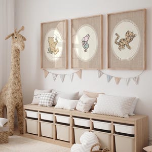 May include: Three framed wall art prints featuring illustrations of Winnie the Pooh, Piglet, and Tigger inside oval borders. The prints are displayed in light wood frames above a wooden storage bench with white and beige bins and decorative bunting.