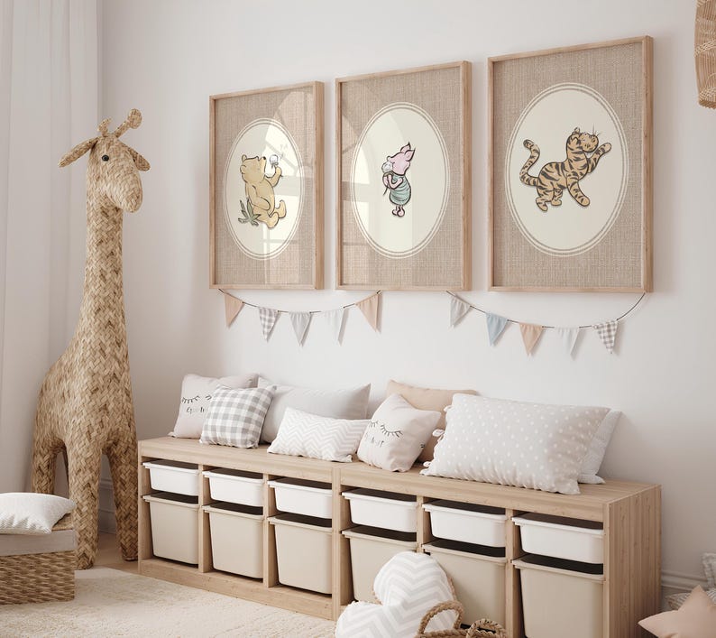 May include: Three framed wall art prints featuring illustrations of Winnie the Pooh, Piglet, and Tigger inside oval borders. The prints are displayed in light wood frames above a wooden storage bench with white and beige bins and decorative bunting.