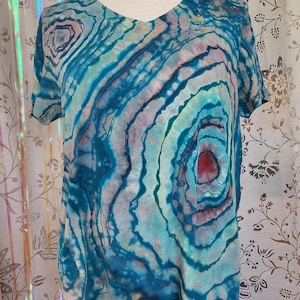 Pode incluir: Uma camiseta com decote em V da Lands' End Canvas com um padrão tie-dye único. A camisa apresenta círculos concêntricos em tons de azul, turquesa e roxo, com um centro vermelho. O design geral assemelha-se a uma pedra de ágata.