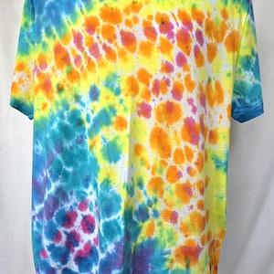Könnte beinhalten: Ein kurzärmeliges T-Shirt mit Batikmuster in einer lebendigen Farbpalette. Das Shirt weist eine Mischung aus Blau-, Gelb-, Orange- und Lilatönen auf, die ein einzigartiges und auffälliges Muster erzeugen. Das Shirt wird auf einer weißen Schaufensterpuppe präsentiert.