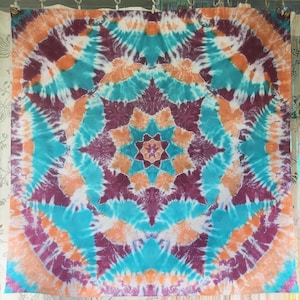 May include: A square, tie-dye textile with a symmetrical design. The pattern features concentric circles and starbursts in shades of teal, orange, purple, and white. The textile is likely a tapestry or wall hanging.
