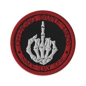 May include: Embroidered patch with a black center and a red border. The patch features a white skeleton hand giving the middle finger. The text "OFFENDING SIMPLE MINDS SINCE DAY ONE" is embroidered around the edge.