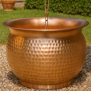 May include: A copper-colored, hammered-finish garden water basin with a decorative chain. The basin is round with a wide rim and sits on a bed of small stones. A green-patina chain hangs from above.