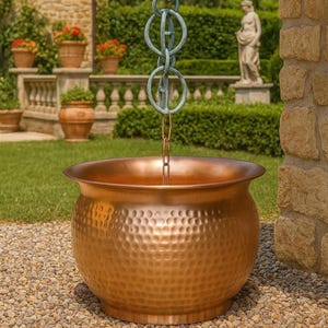 May include: A round, hammered copper planter bowl suspended by a vertical chain featuring large, weathered teal-colored metal rings at the top. The bowl has a wide, smooth rim and a textured, dimpled surface that reflects light across its metallic body.