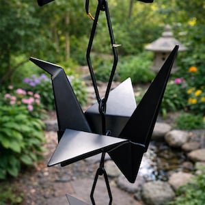 May include: A matte black metal rain chain featuring stylised origami crane shapes connected by diamond-shaped links. The structure is composed of rigid geometric bird forms with pointed wings and tails, suspended vertically in a repeating pattern.