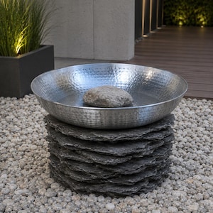 May include: A circular, silver-toned metal bowl with a hammered texture, containing a single grey stone. The bowl is supported by a cylindrical base constructed from stacked, dark grey, rough-edged slate pieces, creating a tiered, natural appearance.
