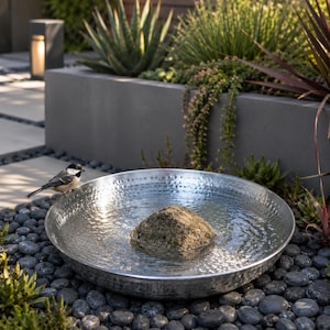 May include: A round, silver-colored metal birdbath with a hammered texture, filled with water and containing a single, rough-textured, tan-colored stone in the center. A small black, white, and gray bird is perched on the rim of the basin.