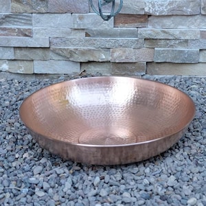 May include: A large, hammered copper bowl with a wide, shallow design. The bowl has a rich, reddish-brown color and a textured surface. It sits on a bed of small, gray stones, with a stone wall in the background. The bowl is likely for decorative or functional use.