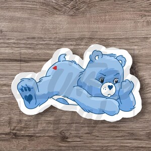 May include: A light blue sticker featuring a cartoon bear lying down. The bear has a heart on its belly and on its rear. The bear's face has a heart-shaped nose and a sad expression. The sticker is on a white background.