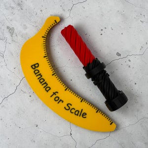 May include: A yellow banana-shaped ruler with the text "Banana for Scale" in black. A red and black object with a textured design is next to the ruler. The ruler has a measurement scale along the curved edge.