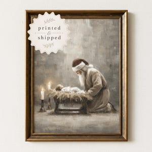 May include: A framed oil painting print featuring a bearded figure in a Santa hat kneeling beside a baby lying in a manger. Two lit candles stand nearby. The artwork has a muted, textured gray and brown color palette. Text reads printed & shipped.