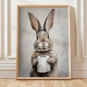 Puede incluir: Una impresión artística enmarcada que muestra un conejo marrón sosteniendo un rollo de papel higiénico. El conejo está pintado con detalles de pelaje y ojos grandes. La obra de arte está enmarcada en madera clara, sobre un fondo gris neutro.